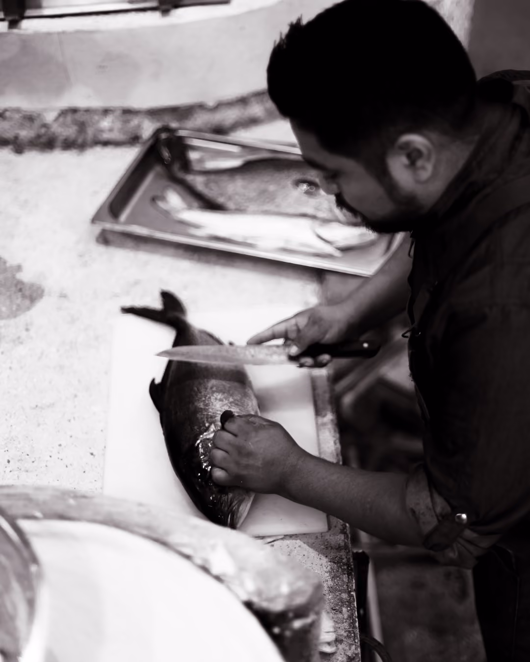 Man filleting a large fish on a cutting board with a knife in a kitchen setting, with another fish on a metal tray in the background.