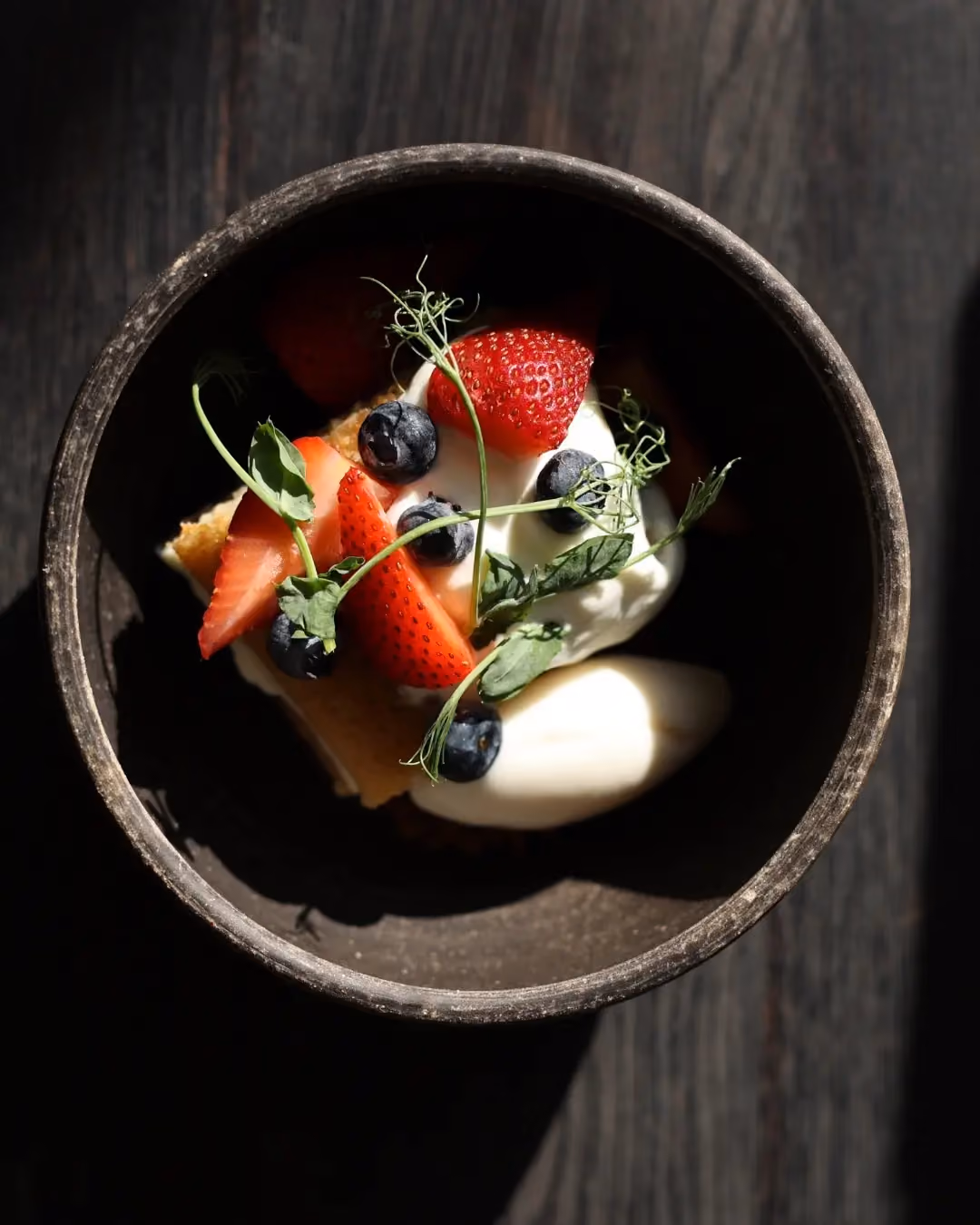 Top view of a dessert in a dark ceramic bowl on a wooden surface, featuring a slice of cake topped with whipped cream, fresh strawberries, blueberries, and green herb garnishes.
