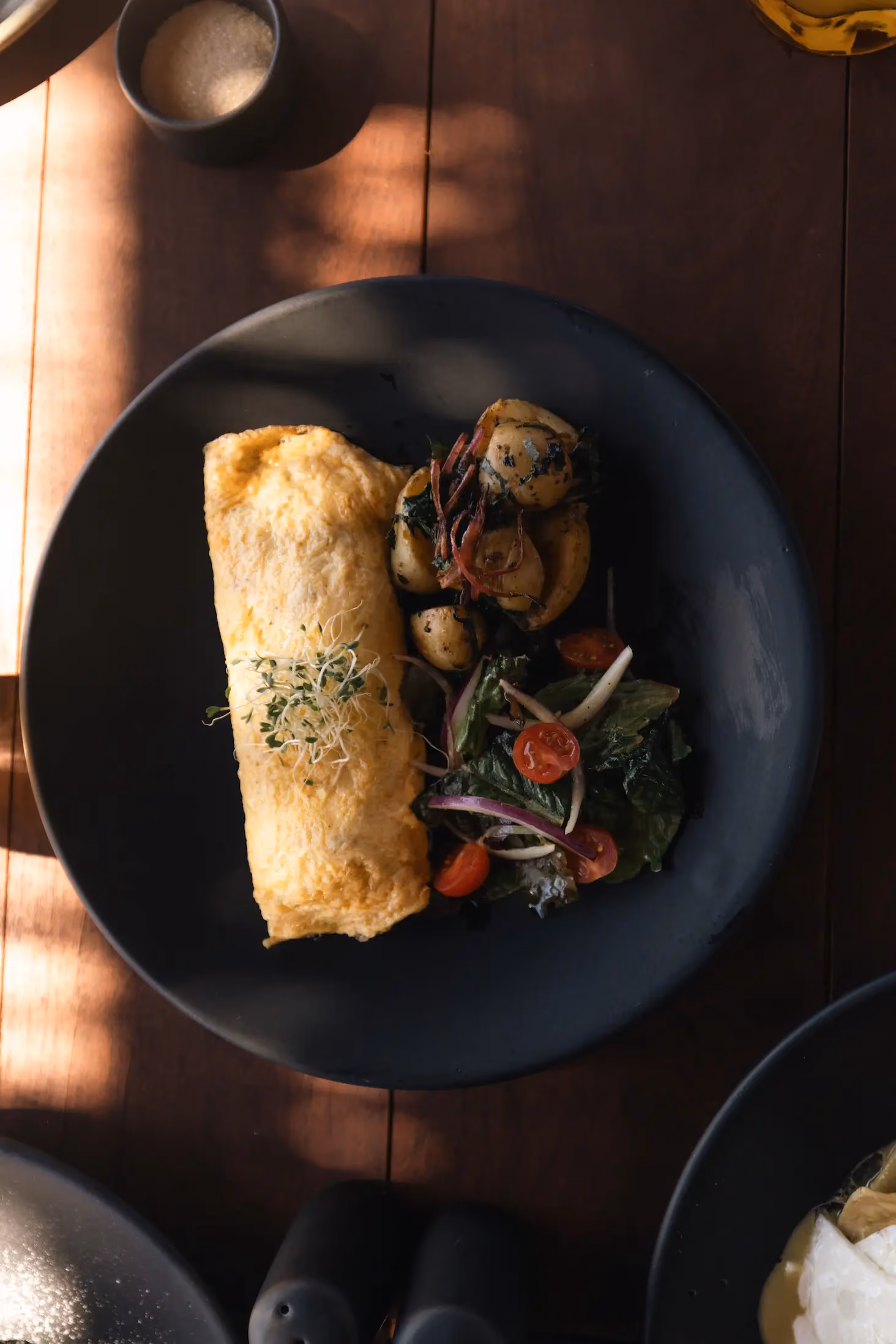 Black plate with an omelet topped with sprouts, served with roasted potatoes and a side salad of cherry tomatoes, leafy greens, and sliced onions on a wooden table with soft lighting.