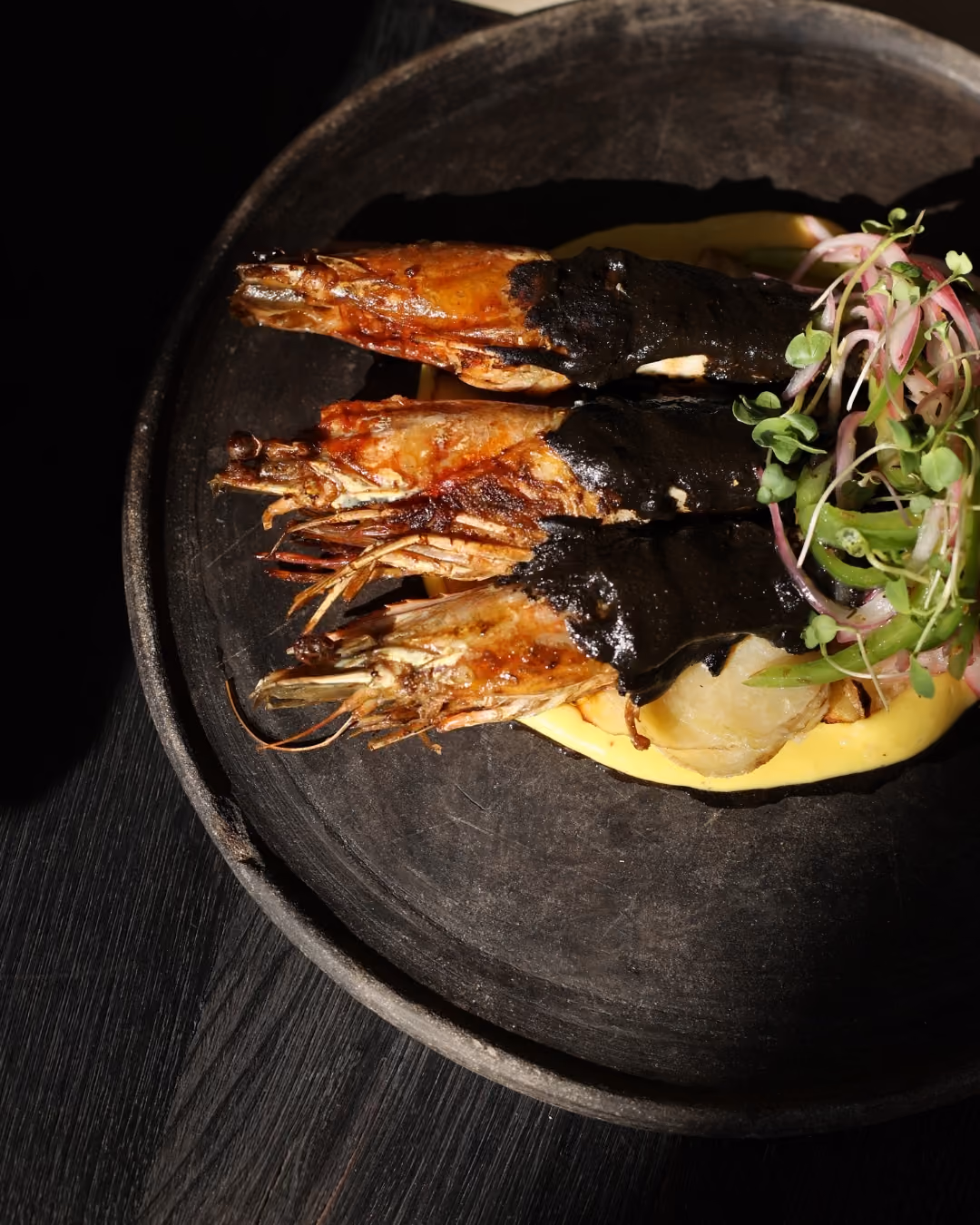 Three cooked prawns partially covered with a black sauce, placed on a bed of yellow sauce with some sliced vegetables and microgreens on a dark round plate.