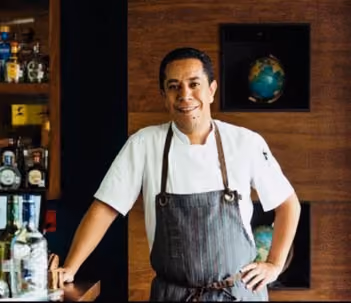 Smiling chef wearing a white shirt and striped apron, standing with one hand on a counter and the other on his hip in a bar area with bottles and a globe in the background.