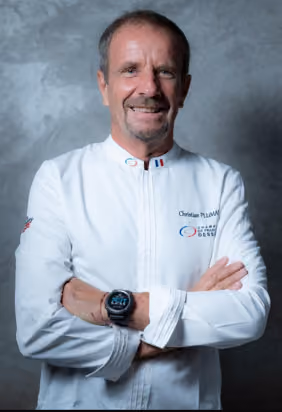 Smiling middle-aged man with short hair and a goatee wearing a white chef's jacket embroidered with the name Christian Plumier and logos, arms crossed, against a gray textured background.