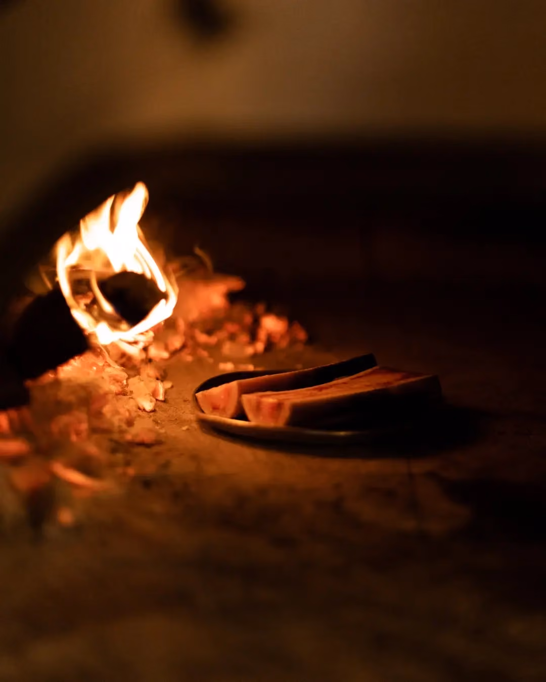Two bone marrow pieces on a plate placed near a wood fire with glowing embers inside a rustic oven.