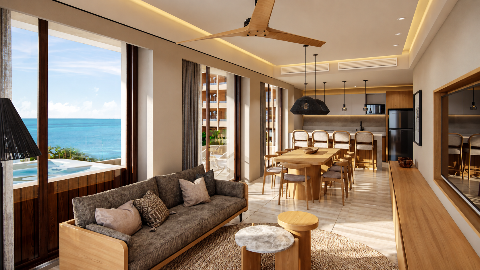 Modern living room and dining area with wooden furniture, gray sofa with cushions, round nesting tables, large windows showing ocean view, ceiling fan, pendant lights above dining table, and a kitchen with bar stools and stainless steel appliances.