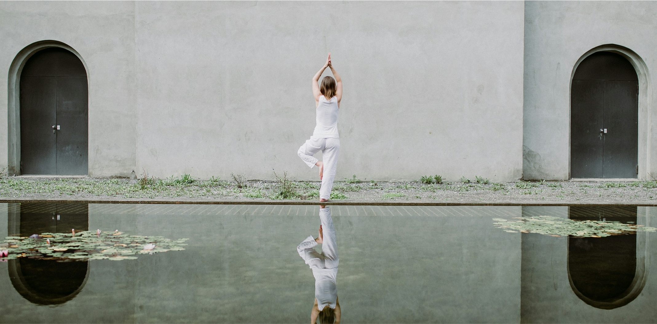 Person in white clothing practicing yoga in tree pose in front of a concrete wall with two arched doors and a reflective pond with lily pads.