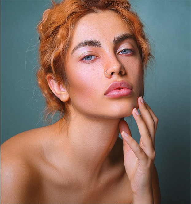 Close-up portrait of a young woman with red hair, blue eyes, and a nose ring, resting her hand on her chin.