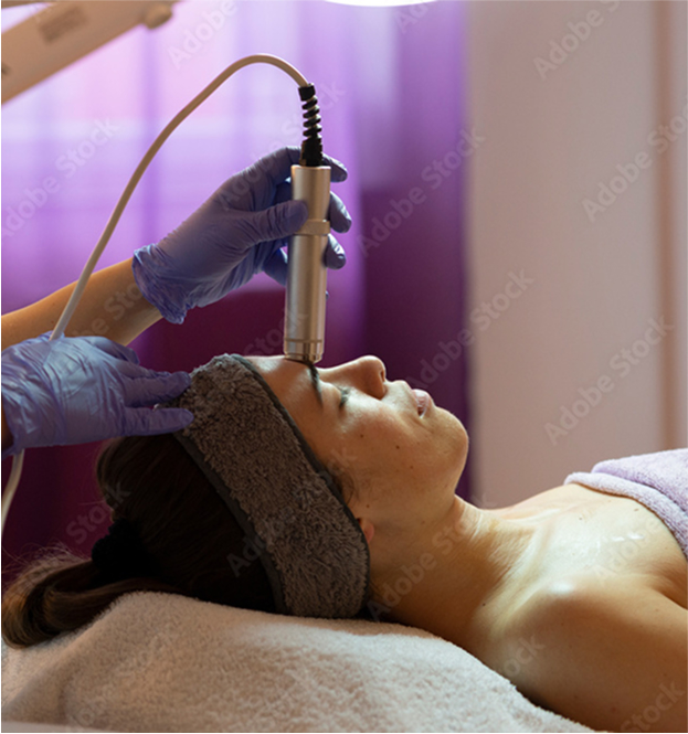 Side view of a woman receiving a facial treatment with a handheld device by a gloved professional in a spa setting.