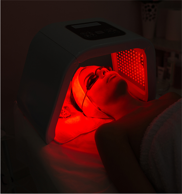 Person wearing protective goggles receiving red light therapy on their face under a specialized device.