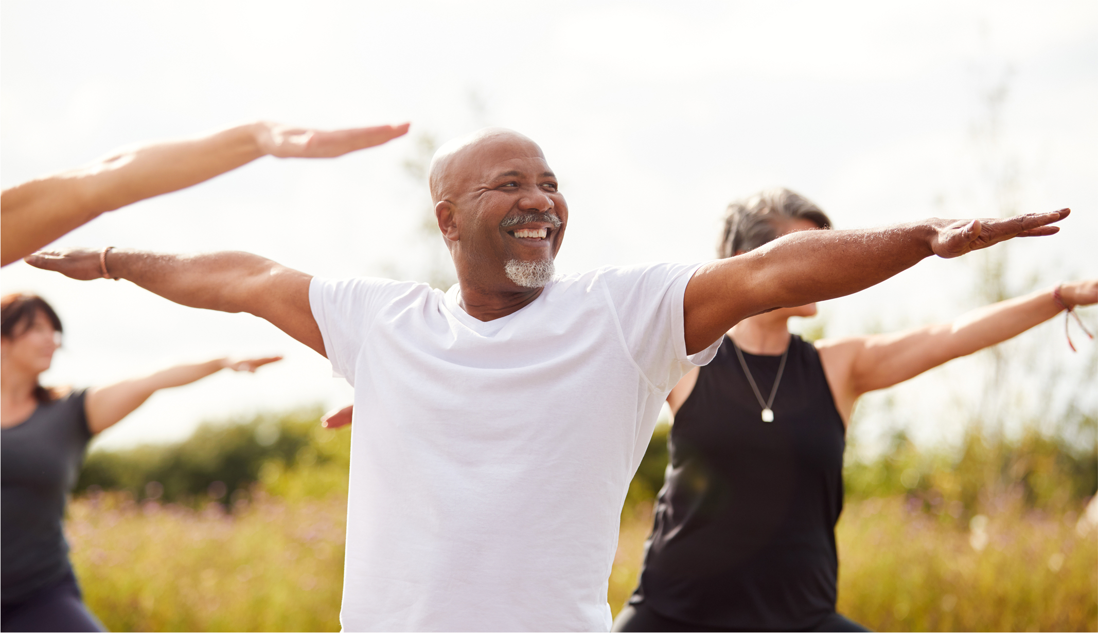 Group of people outdoors practicing yoga, with a smiling man in white shirt extending his arms.