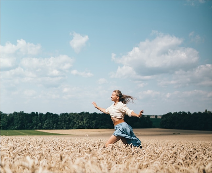 Woman running joyfully through a golden wheat field under a blue sky with scattered clouds.