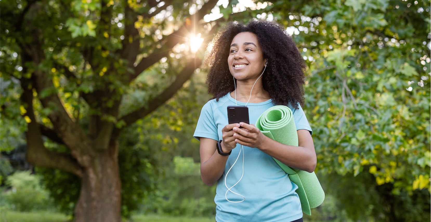 Smiling woman with curly hair holding a green yoga mat and smartphone outdoors with sun shining through trees.