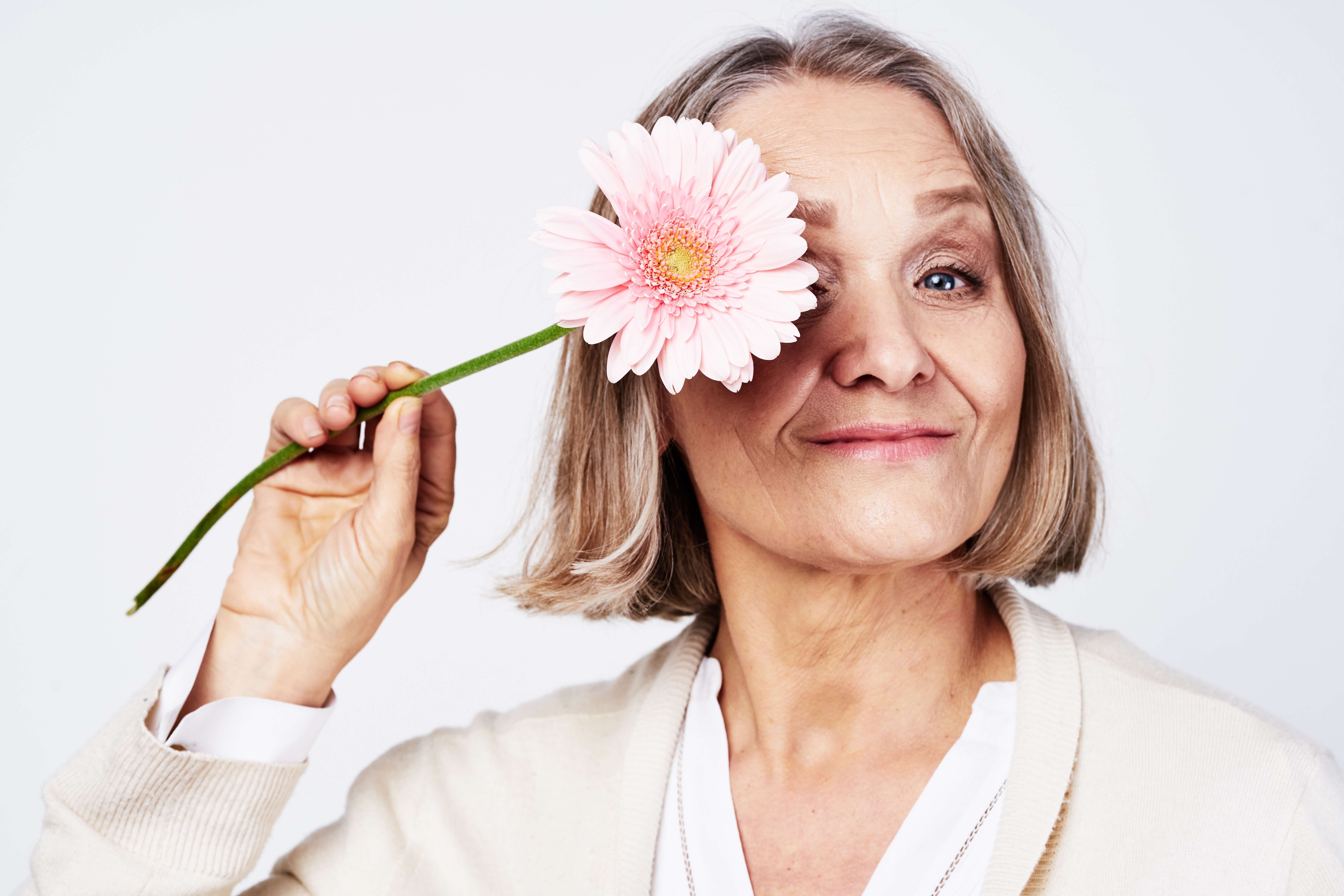 Smiling middle-aged woman holding a pink gerbera flower covering one eye against a light background.