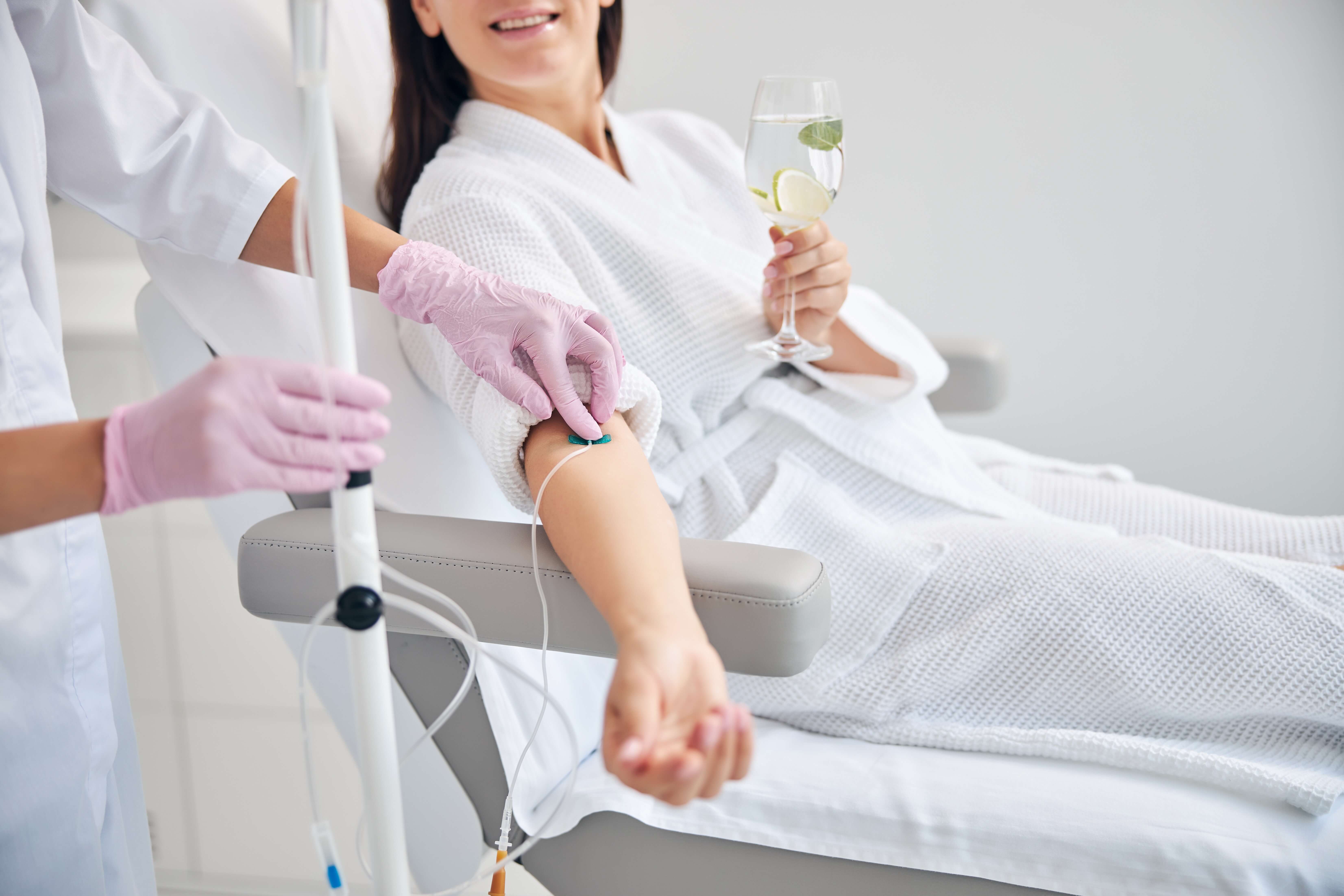 Woman in white robe receiving IV treatment while holding a glass of water with lemon and mint.