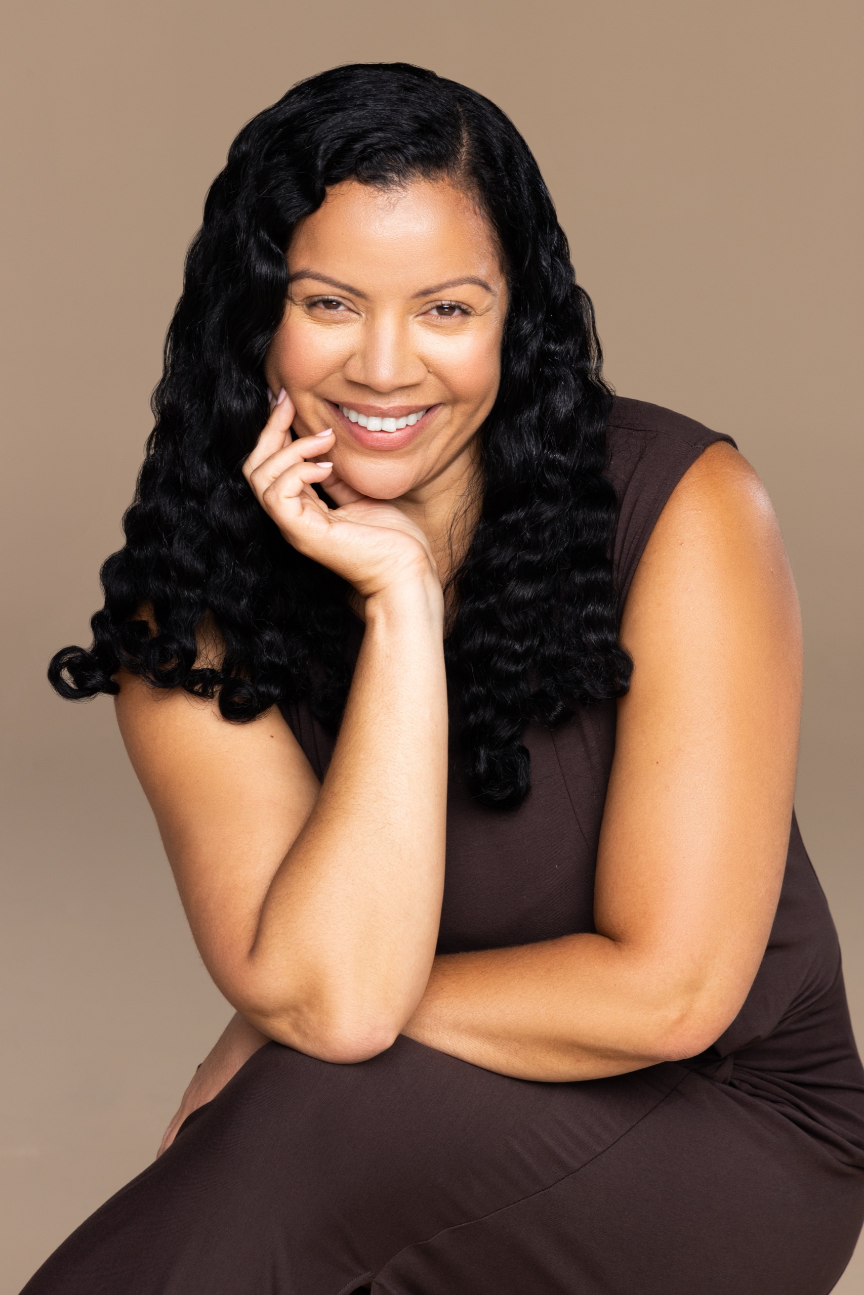 Smiling woman with long curly black hair resting her chin on her hand against a beige background.