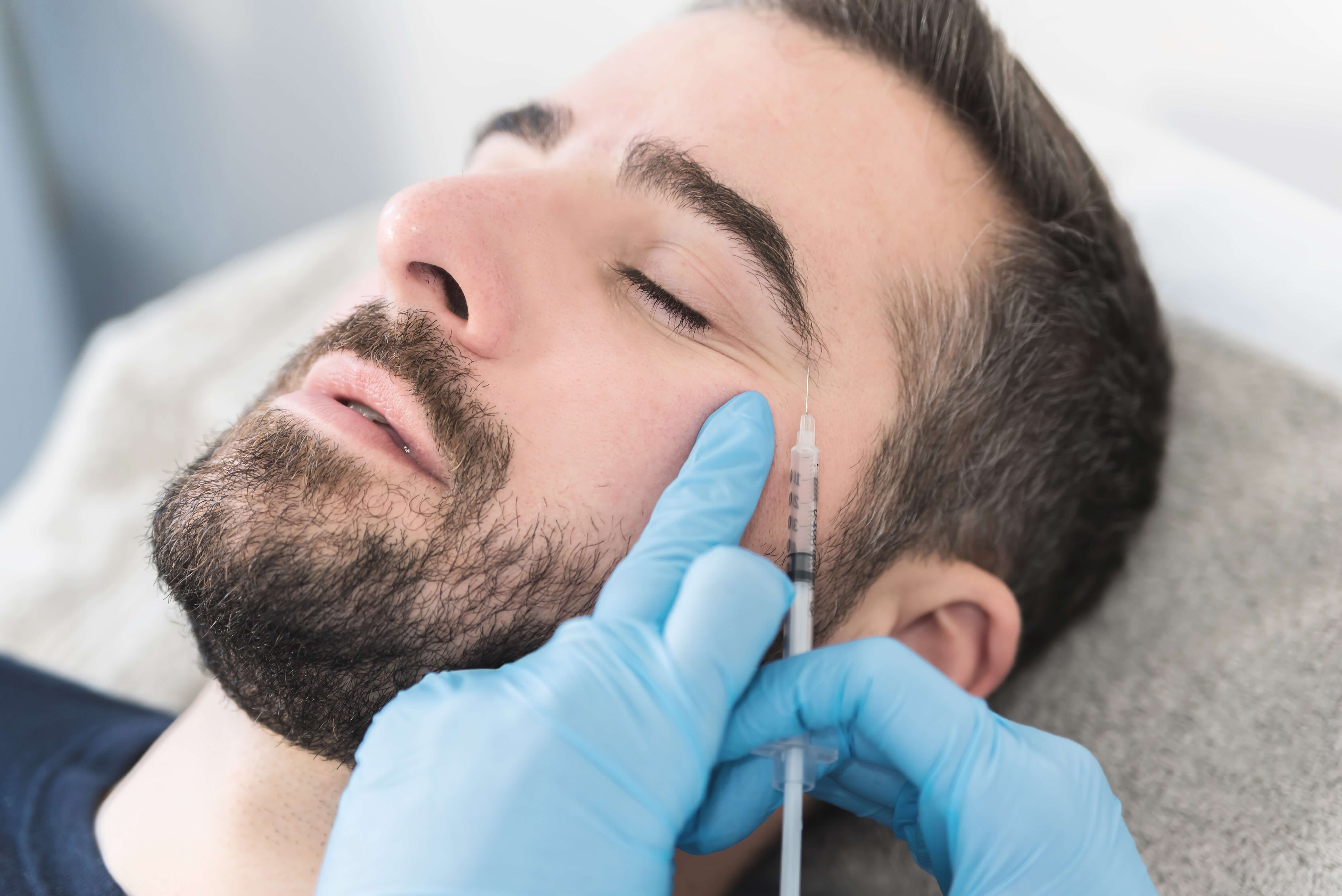 Close-up of a man receiving a cosmetic injection near his closed eye by a gloved hand holding a syringe.