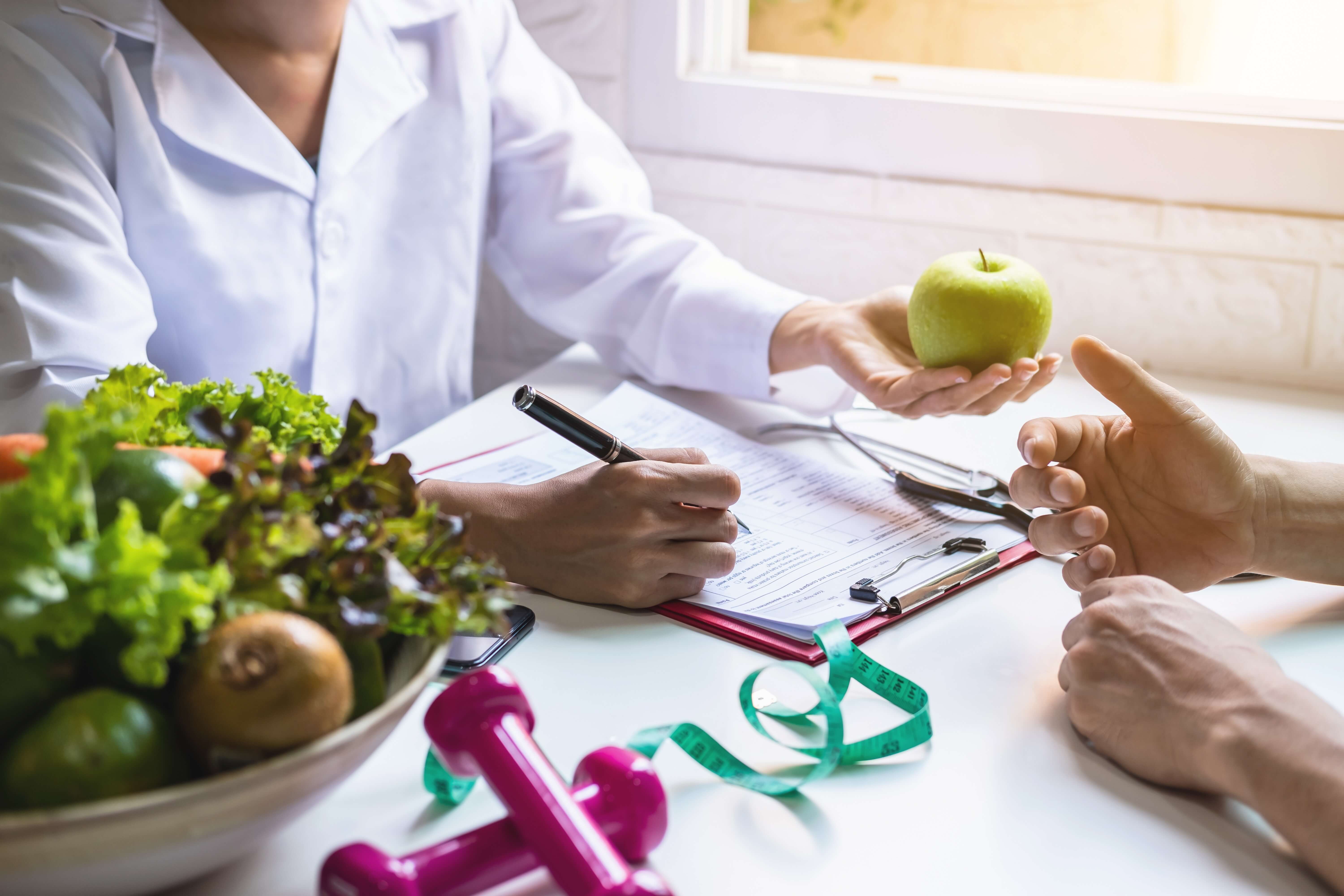 Nutritionist offering a green apple to a person while taking notes on a clipboard, with fresh vegetables, pink dumbbells, and a measuring tape on the table.