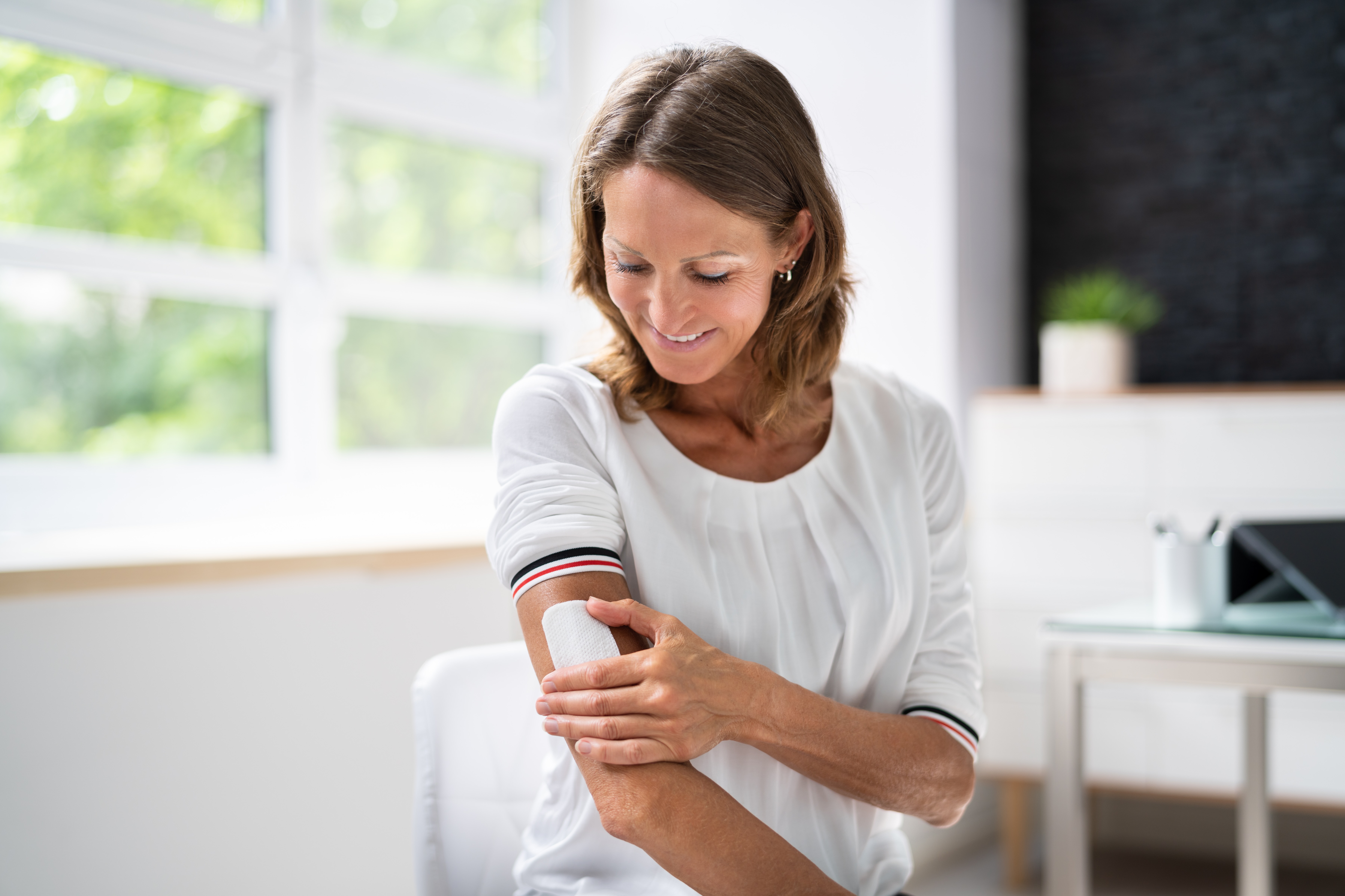 Smiling woman in a white shirt applying a bandage on her elbow in a bright room.