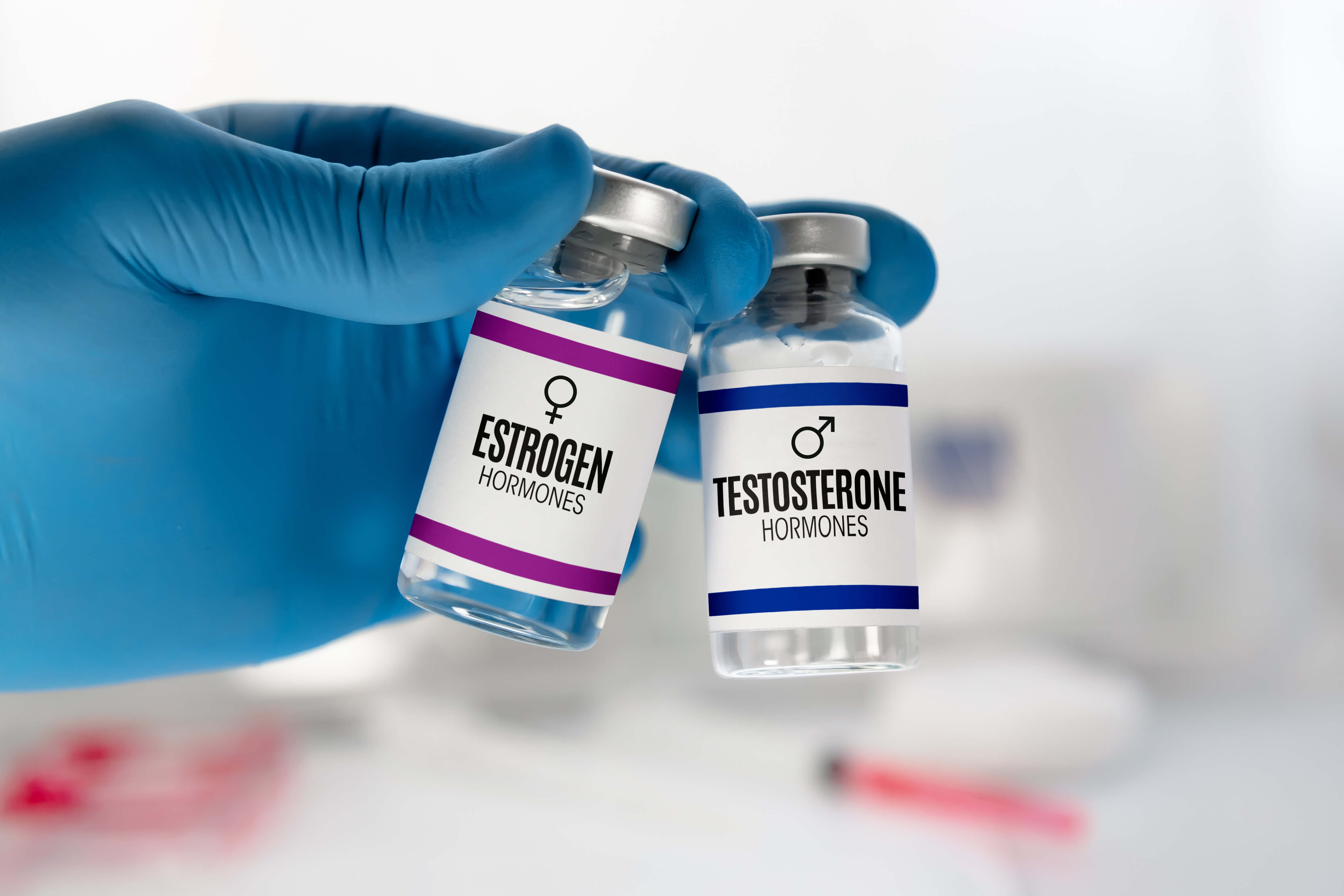 Gloved hand holding two vials labeled Estrogen Hormones and Testosterone Hormones.