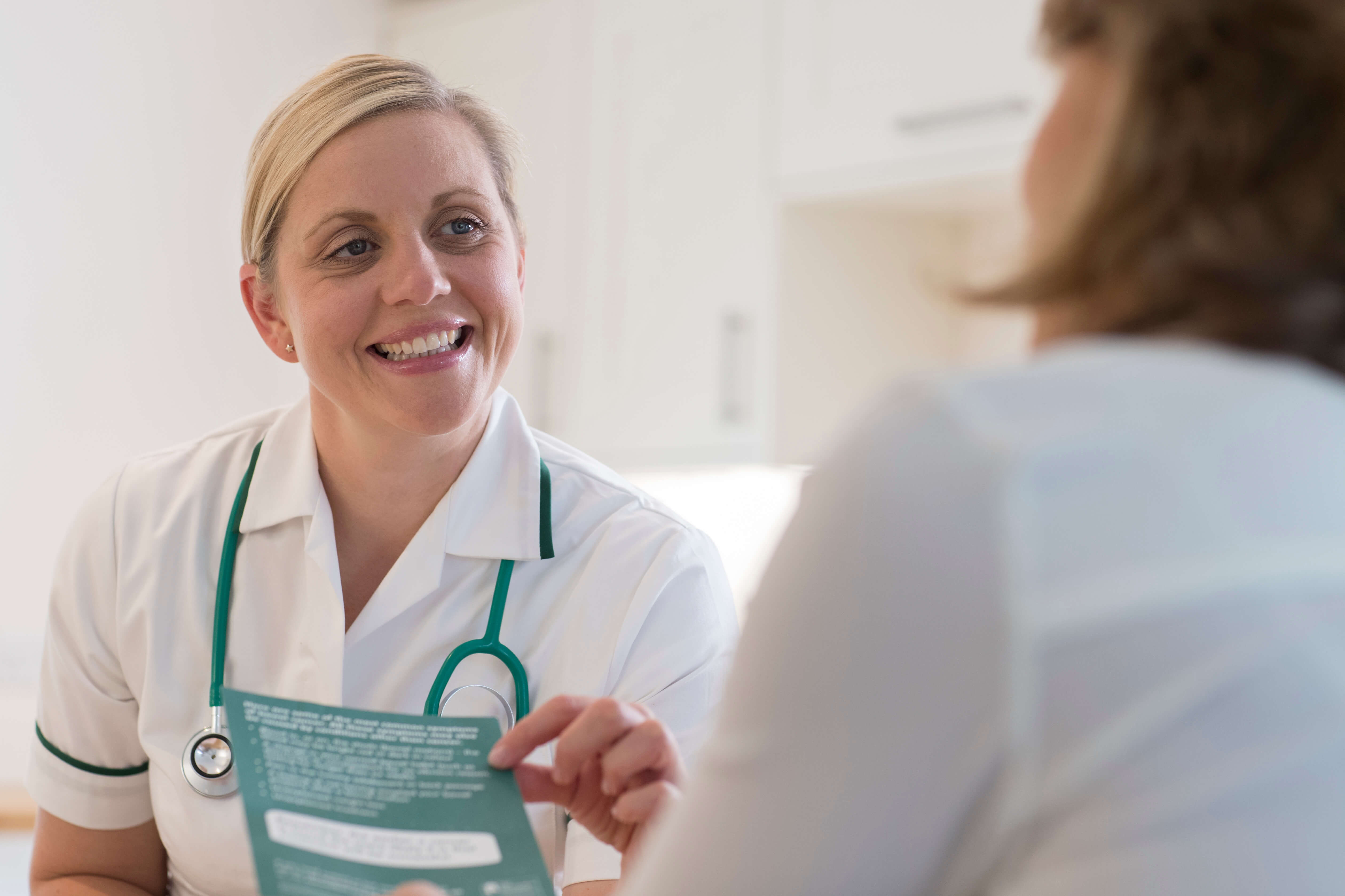 Smiling female nurse with stethoscope speaking to a patient while holding a paper.