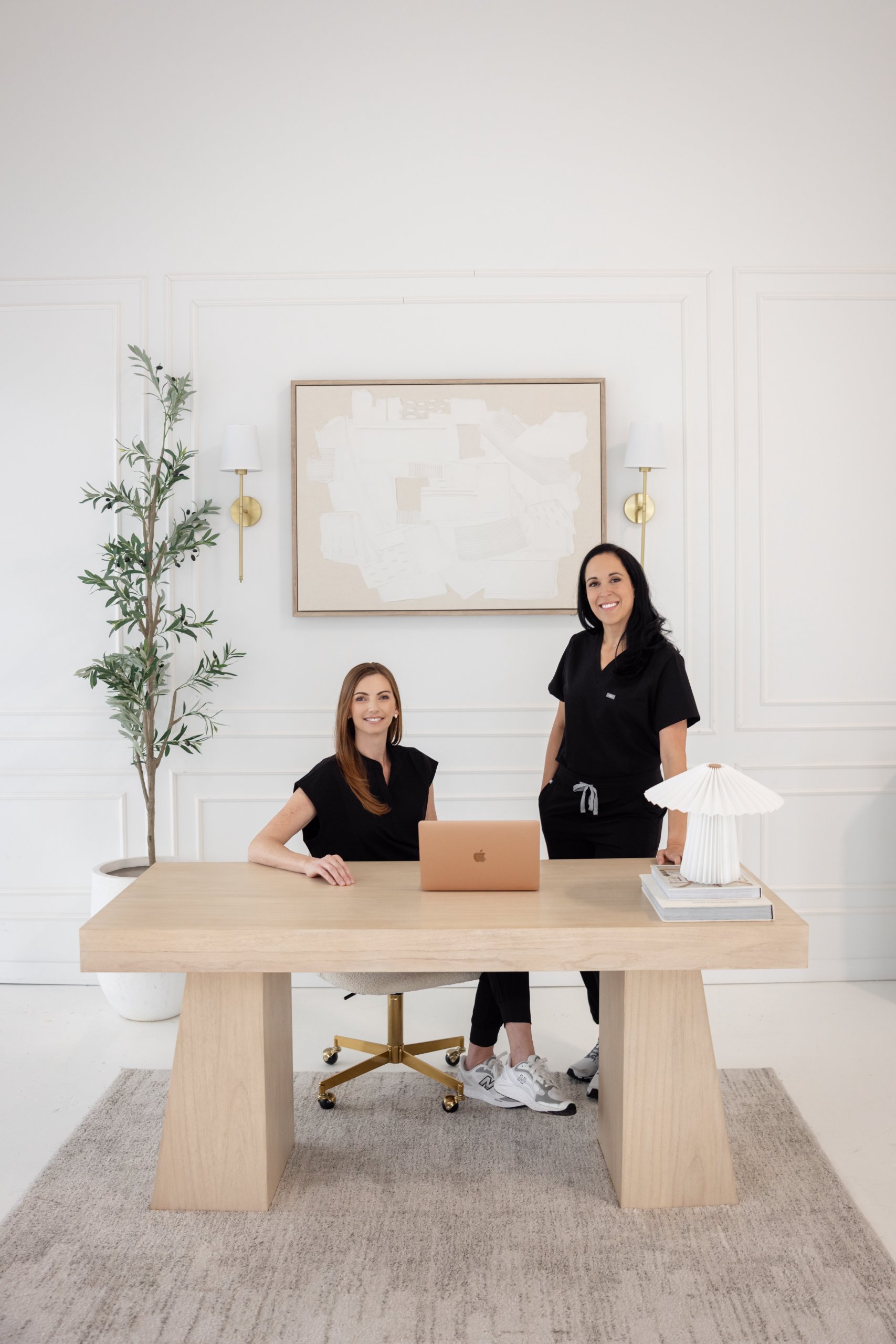 Two women in black outfits posing in a bright, minimalistic office with a wooden desk, laptop, plant, and modern decor.