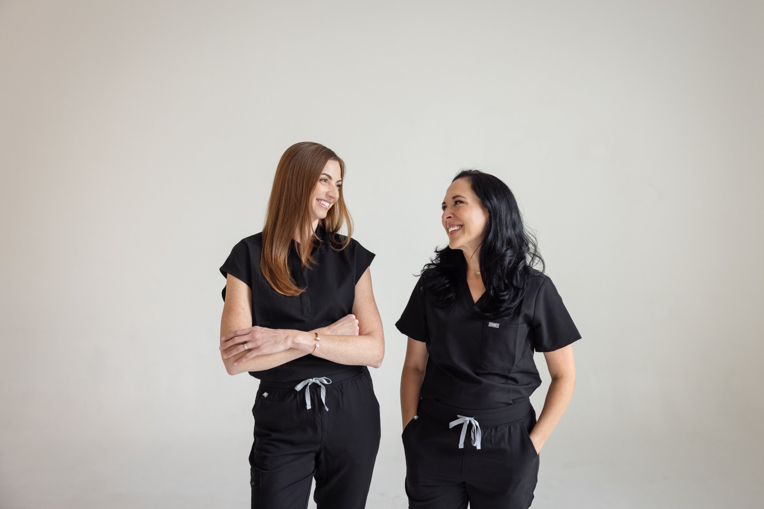Two women smiling and looking at each other, both wearing black medical scrubs with gray drawstrings.