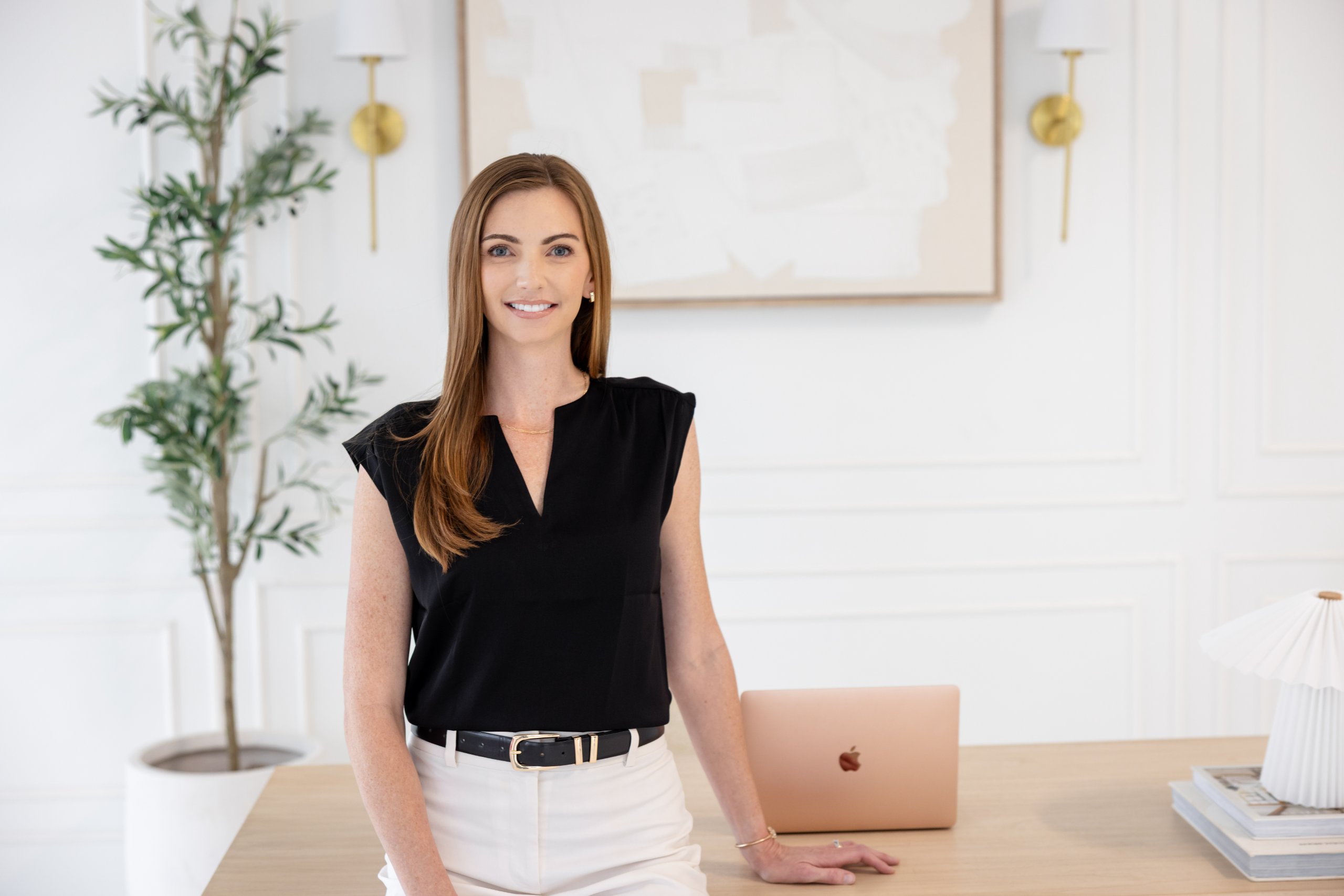 Smiling woman in black sleeveless top and white pants leaning on a desk with a laptop and lamp in a bright office.