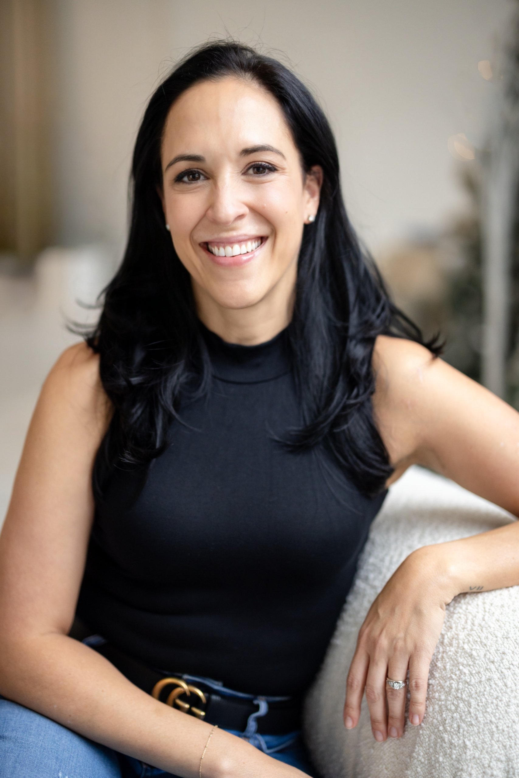 Smiling woman with long black hair in a sleeveless black top and jeans sitting on a light-colored couch.