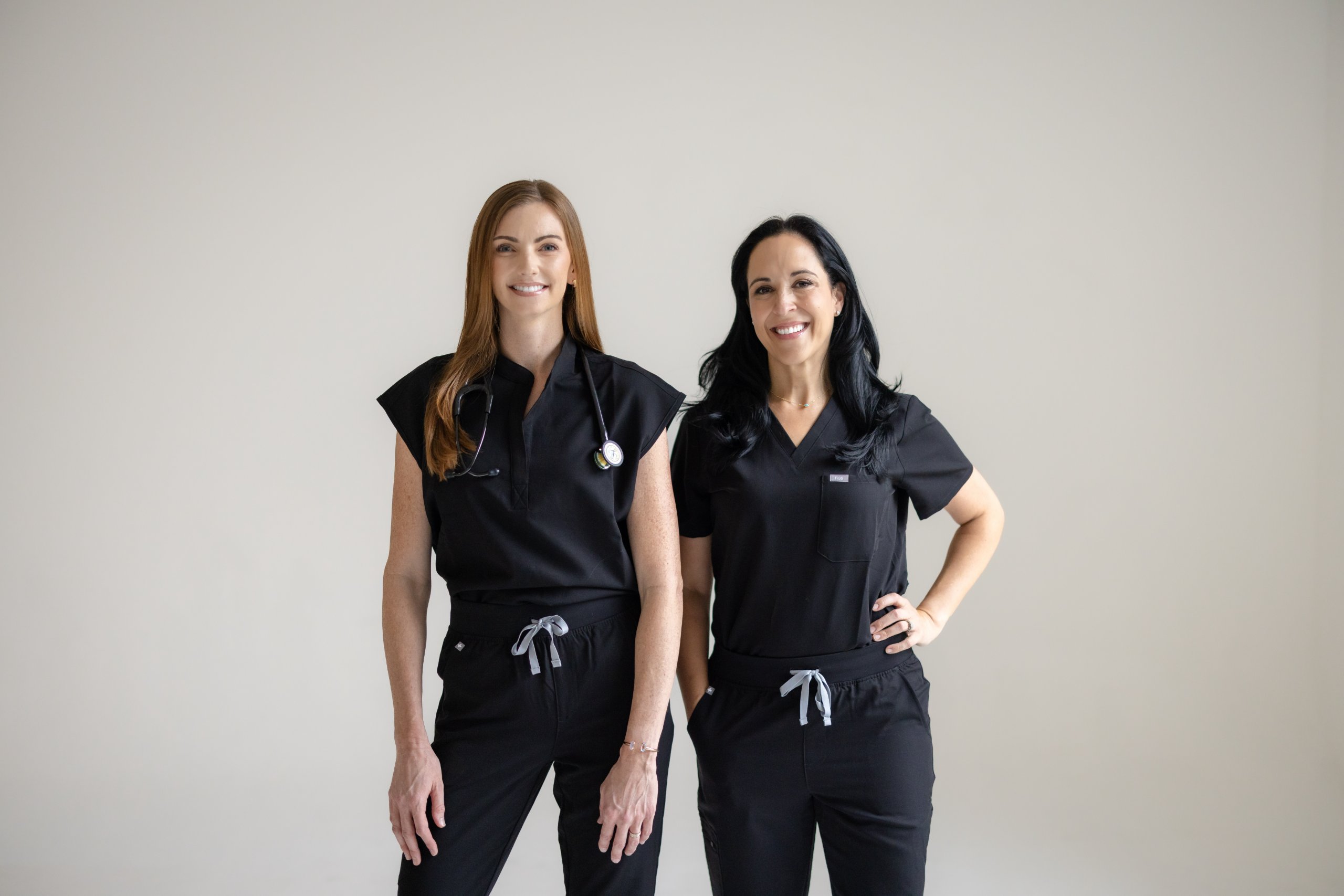 Two smiling female healthcare professionals wearing black scrubs standing against a plain background.
