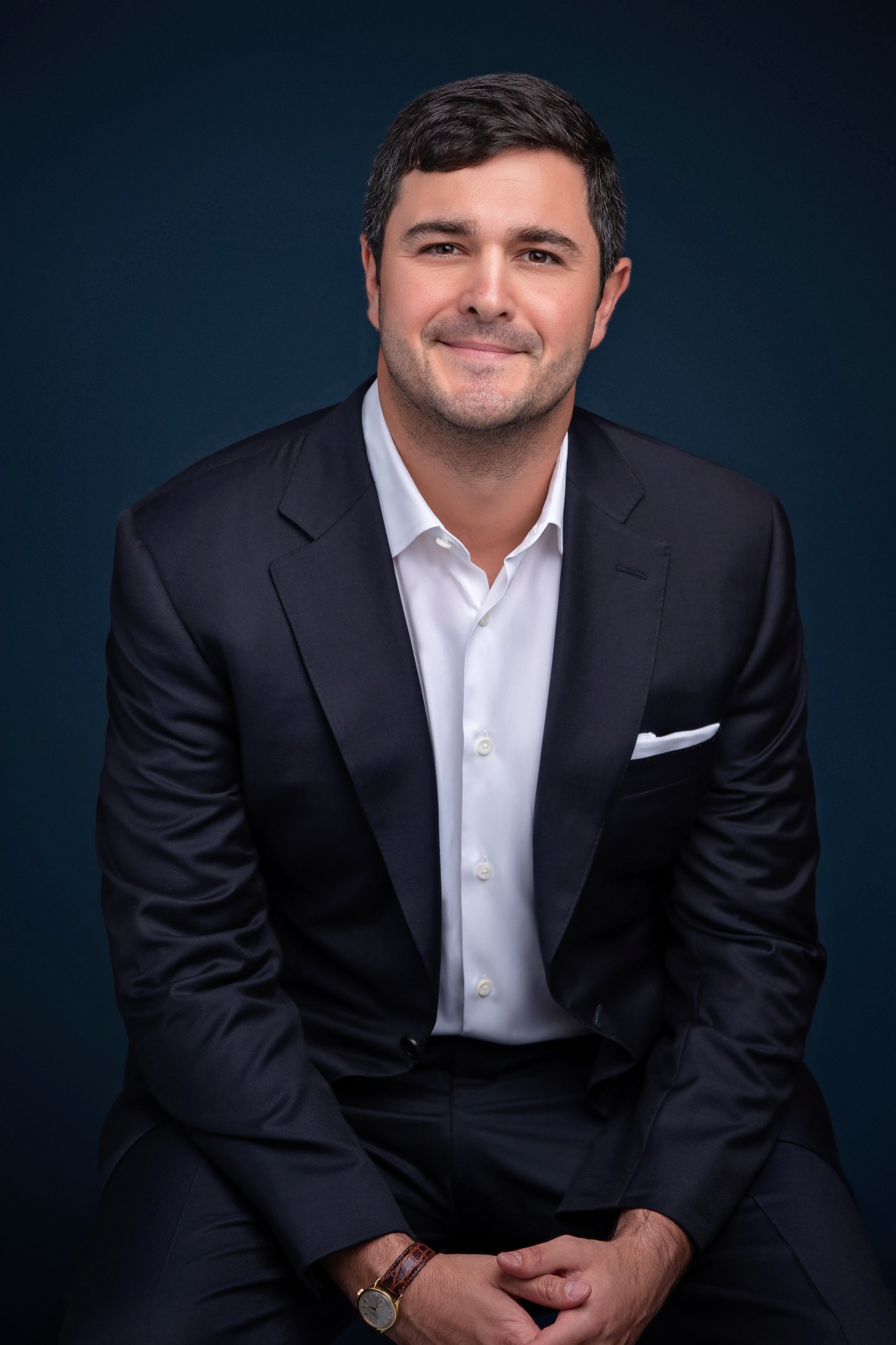 Smiling man in a dark suit and white shirt seated with hands clasped, against a dark blue background.