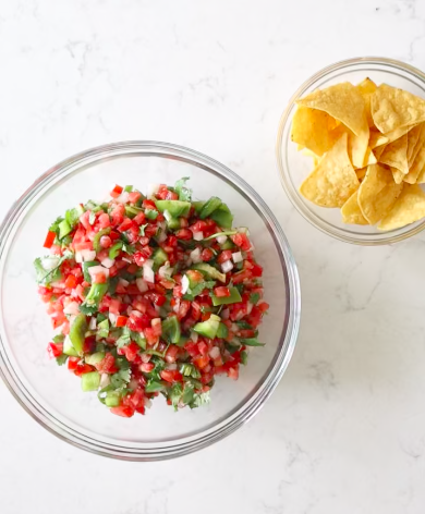 Glass bowl of fresh garden salsa with diced tomatoes, green bell pepper, onion and cilantro, next to a bowl of tortilla chips.