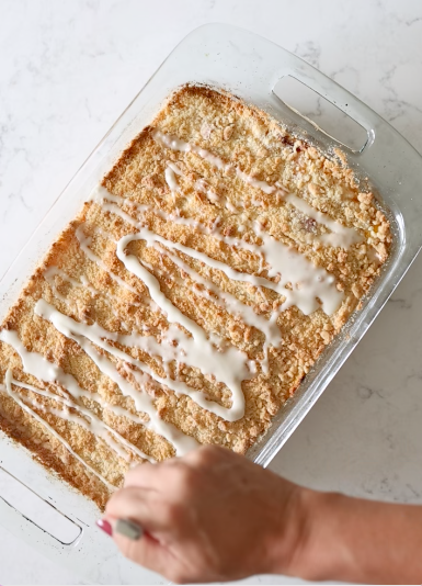 Glass baking dish of golden peach crumb bars being drizzled with white almond glaze.