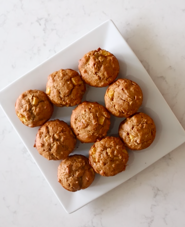 Nine golden pumpkin apple oat muffins flecked with oats and chopped apple, arranged on a square white plate.
