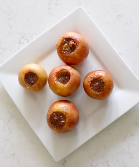 Five baked apples filled with brown sugar and walnuts on a white square plate.