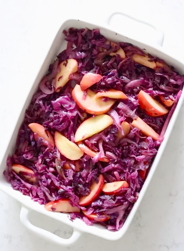 White baking dish of braised red cabbage with sliced onion and wedges of apple stirred through.