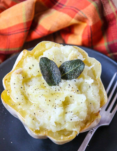 Roasted white acorn squash half filled with creamy mashed squash, topped with cracked black pepper and two crispy fried sage leaves, next to a fork and a plaid napkin.