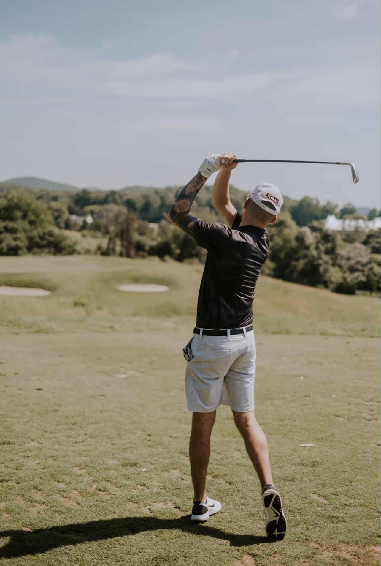 A man in a black shirt and white shorts swings a golf club on a sunny course.