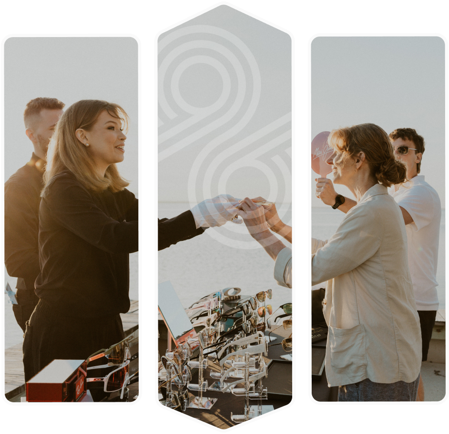 People exchanging handshakes and smiles at an outdoor sunglasses display table, with the ocean in the background.