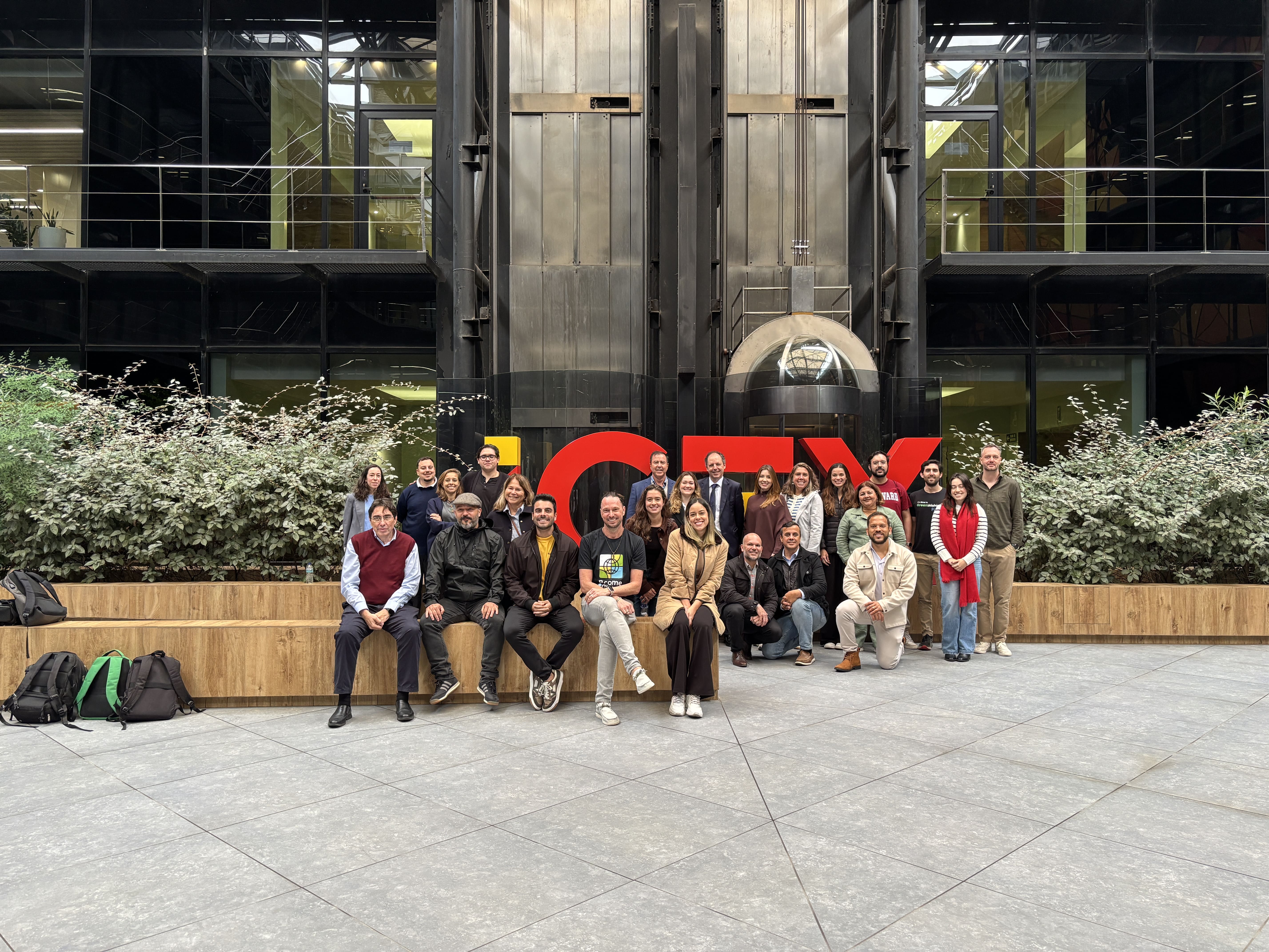 Grande grupo de mais de 20 pessoas posando para uma foto em um pátio externo, algumas sentadas em um muro baixo e outras de pé. Ao fundo, a grande fachada de um prédio em destaque na cor vermelha. As pessoas são participantes e parceiros do Brazilian Impact Techs in Spain, programa que conecta negócios com pessoas e instituições relevantes para a expansão global de impacto.