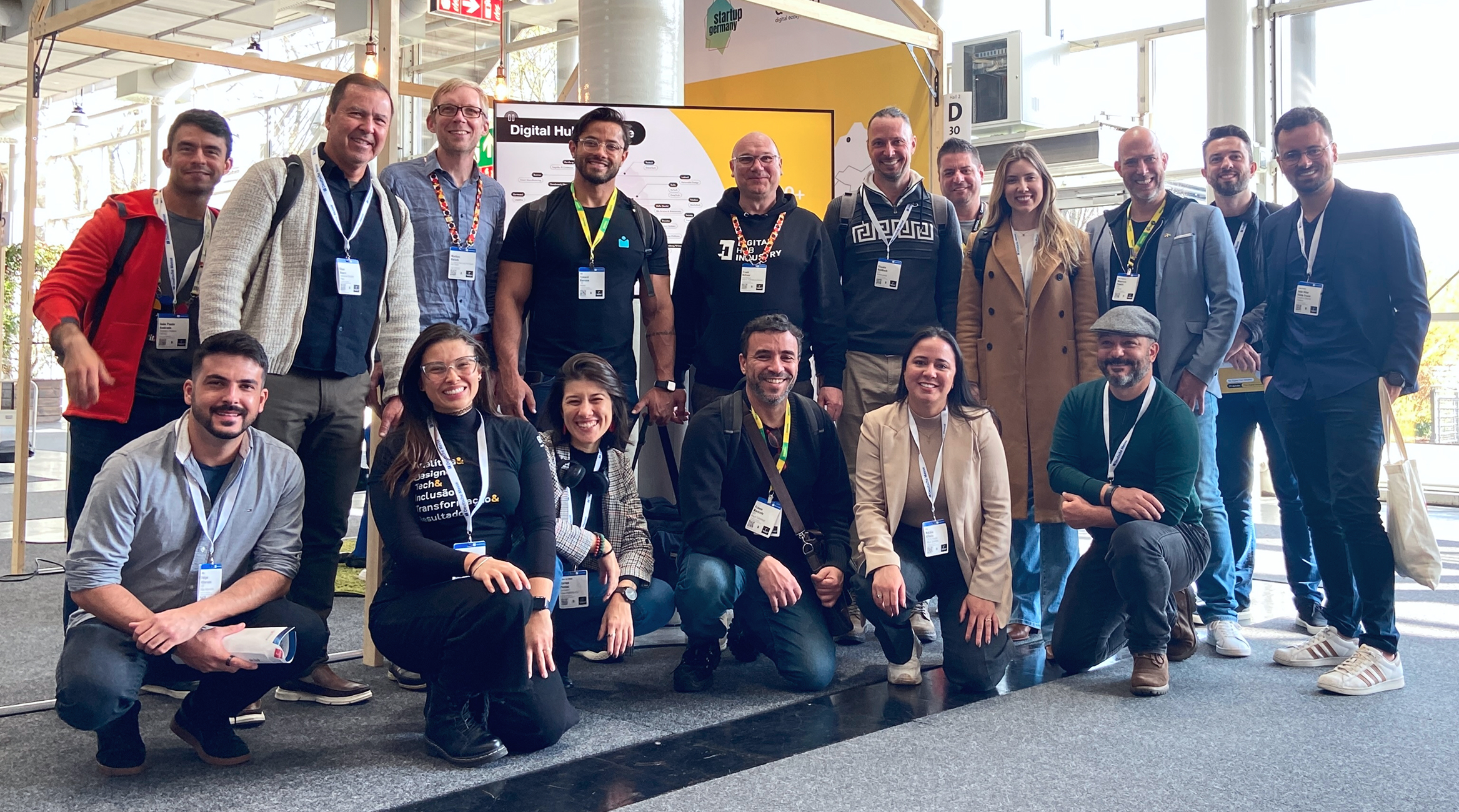 Grupo de 20 pessoas posa para uma foto em um gramado ensolarado em frente a um edifício moderno de vidro. No fundo, à esquerda, há três mastros exibindo a bandeira da Alemanha, a bandeira da União Europeia e um estandarte preto e vermelho com o logotipo da 'Hannover Messe'.