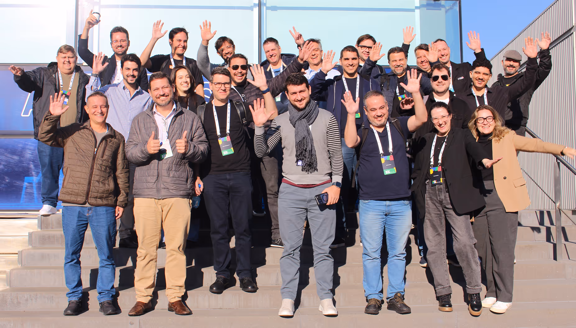 Grupo de 20 pessoas posa para uma foto em um gramado ensolarado em frente a um edifício moderno de vidro. No fundo, à esquerda, há três mastros exibindo a bandeira da Alemanha, a bandeira da União Europeia e um estandarte preto e vermelho com o logotipo da 'Hannover Messe'.