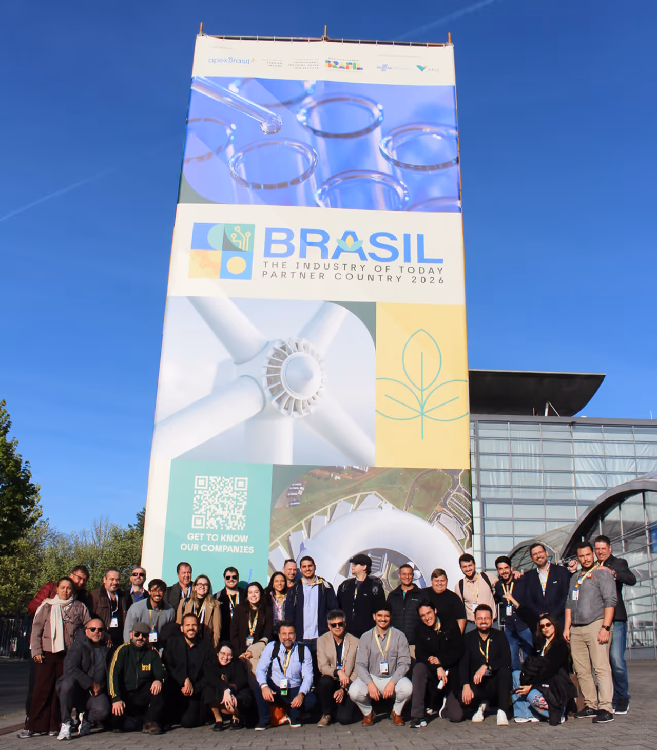 Grupo de 20 pessoas posa para uma foto em um gramado ensolarado em frente a um edifício moderno de vidro. No fundo, à esquerda, há três mastros exibindo a bandeira da Alemanha, a bandeira da União Europeia e um estandarte preto e vermelho com o logotipo da 'Hannover Messe'.