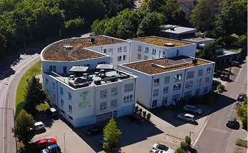 Aerial view of a white U-shaped building with flat roofs and greenery around, surrounded by parked cars and trees.