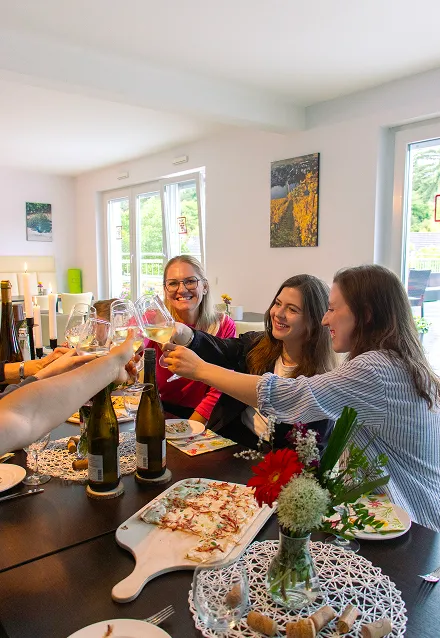 Group of friends clinking wine glasses in a toast around a table with bottles, a flatbread pizza, and a flower vase.