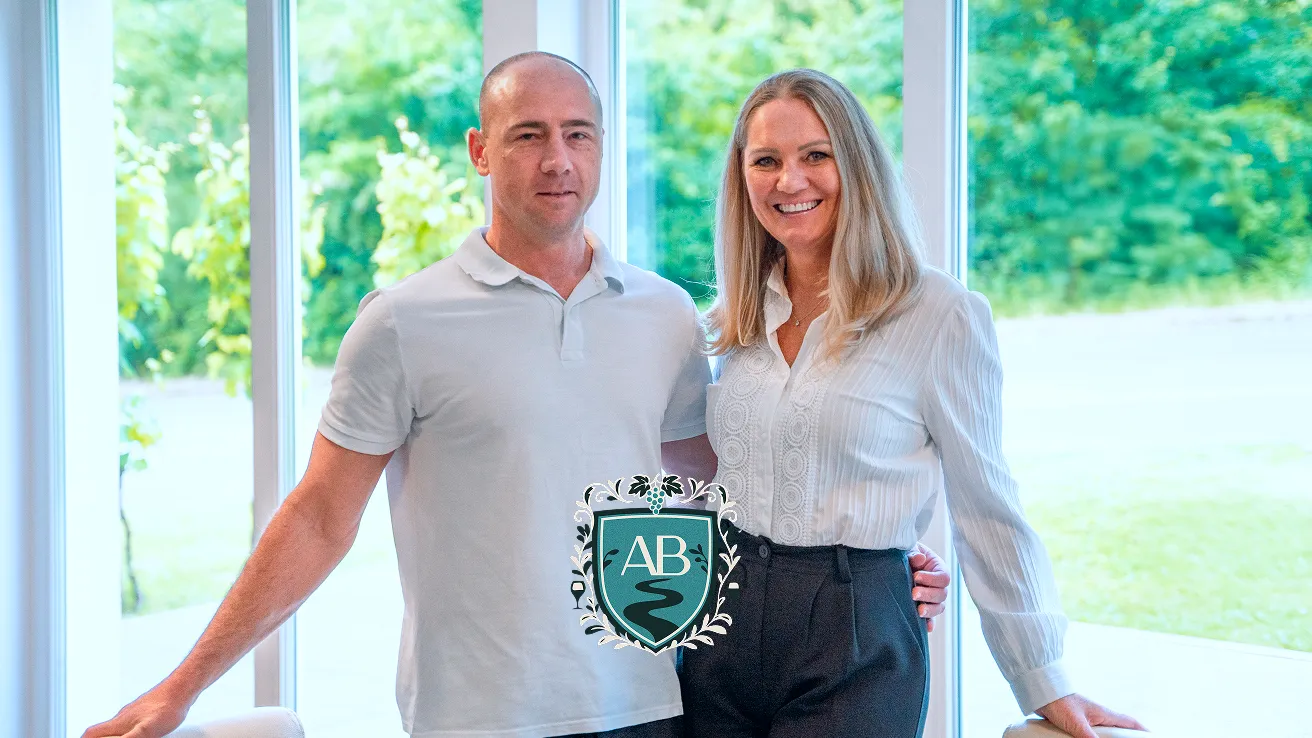 Man in white polo shirt standing next to smiling woman in white blouse and black pants in a bright room with large windows and greenery outside.