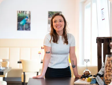 Smiling woman with tattoos leaning on a counter in a bright room with snack jars and framed pictures on the wall.