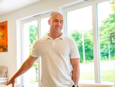 Smiling bald man in a white polo shirt standing indoors near large windows with greenery outside.