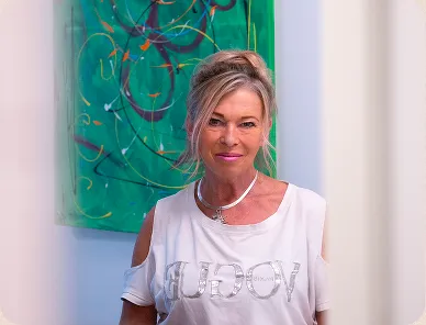 Smiling woman with blonde hair wearing a white shirt with cut-out shoulders and a green abstract painting in the background.