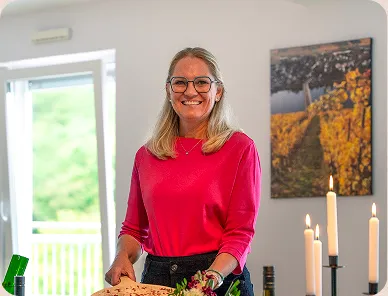 Smiling woman with glasses and blonde hair wearing a pink top holding a dish in a brightly lit room with candles and a framed picture on the wall.