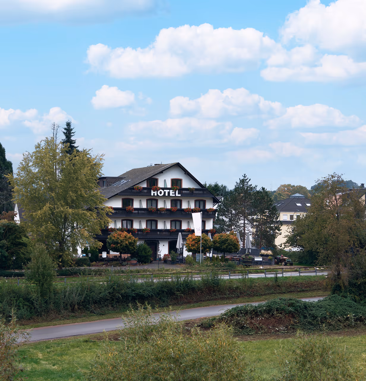 White multi-story hotel with balconies adorned with red flowers, surrounded by trees and greenery under a blue sky with scattered clouds.