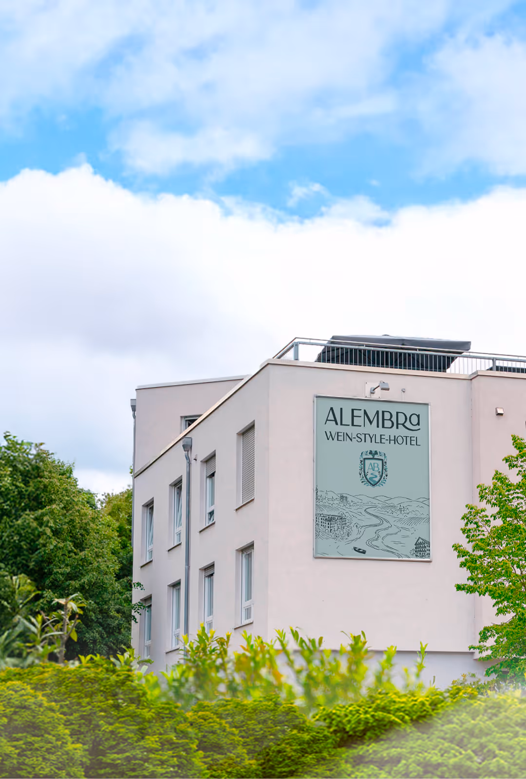 Modern light-colored building with a sign reading 'ALEMBRA WEIN-STYLE-HOTEL' near green trees under a partly cloudy sky.